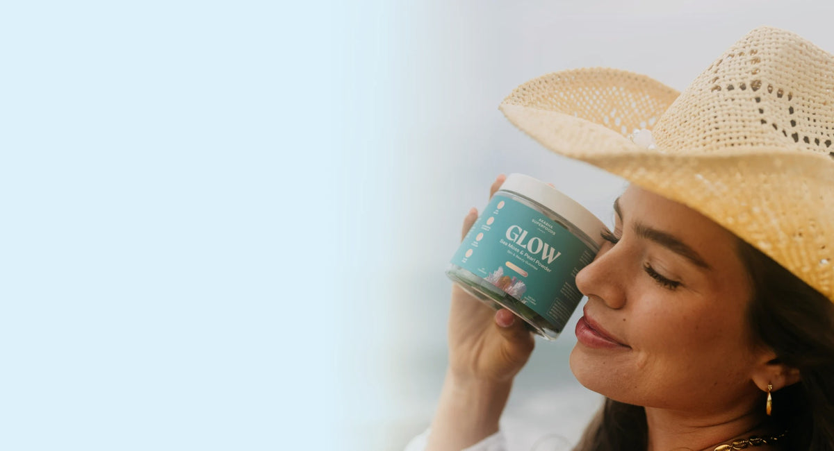 Woman in a straw hat holding a jar of Akasha Sea Moss GLOW Gummies at the beach
