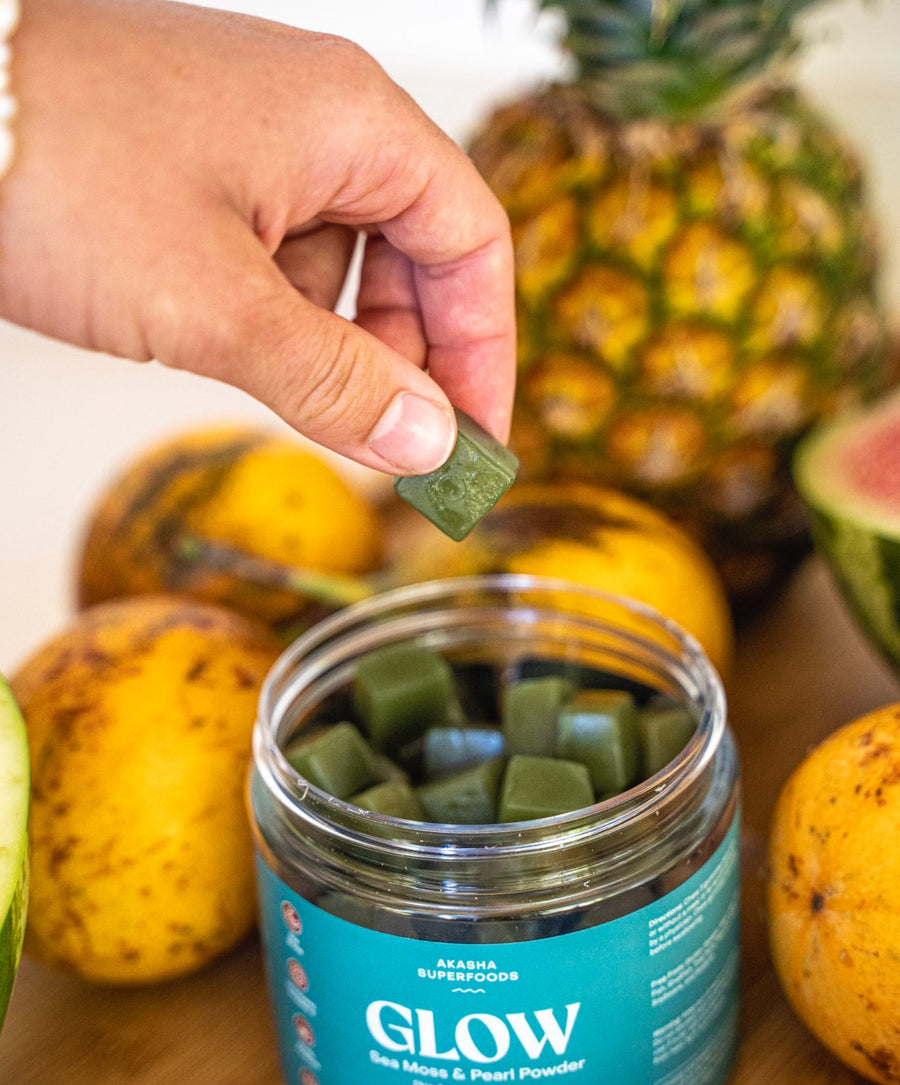 Hand lifting a GLOW gummy from the jar beside fresh pineapple and mango
